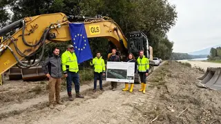 Projektpartner mit EU-Flagge vor Bagger, rechts das Donauufer