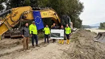 Project partners holding EU-Flagge in front of excavator, danube riverside at the right