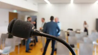 Nahaufnahme Mikrofon, im Hintergrund Besprechungsraum mit Menschen 