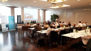 [Translate to English:] Vortrag vor Tischreihen mit etwa zwanzig Zuhörern im Bild